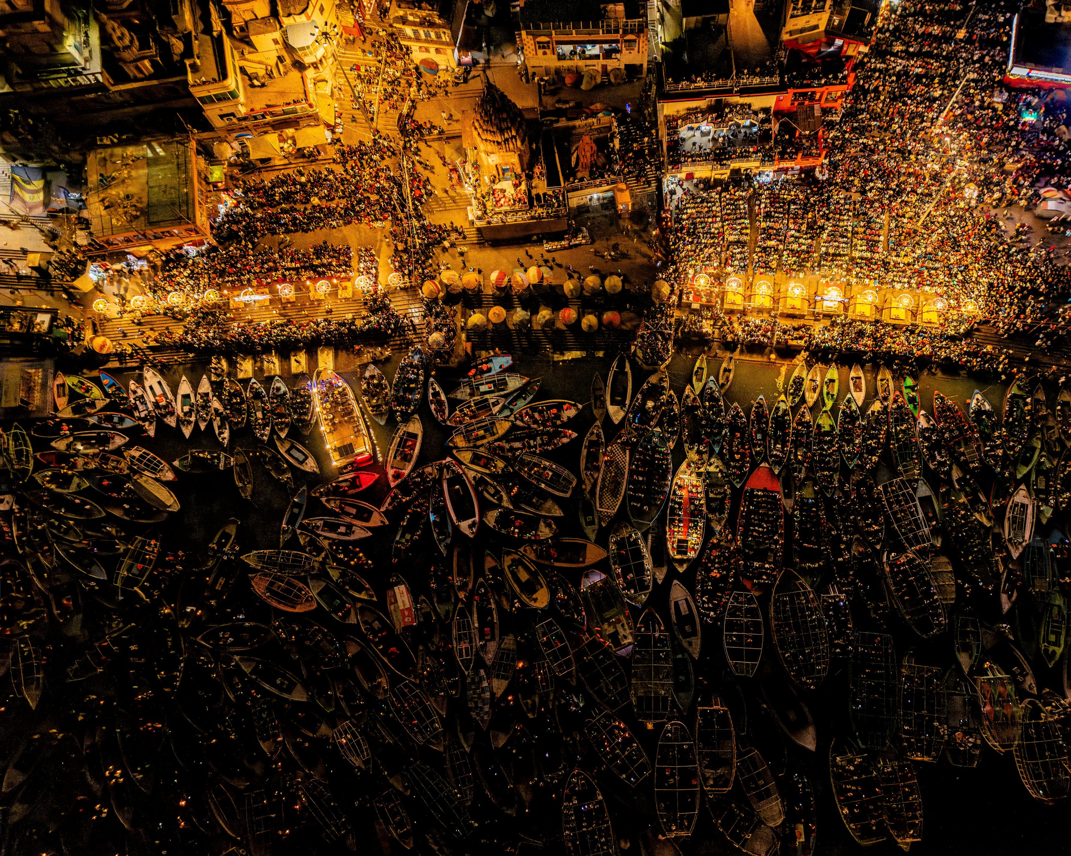 An aerial view of a riverbank packed with people, on shore and in small boats near the shore, seen at night, in India