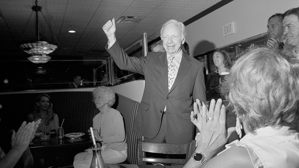 A black-and-white photo of Joe Lieberman in diner wearing a suit and giving a thumbs-up while people gathered around him clap