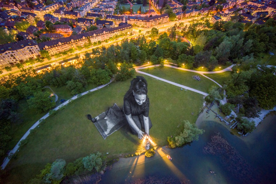 A drone view of a giant girl painted onto grass in a park near brightly lit streets and buildings
