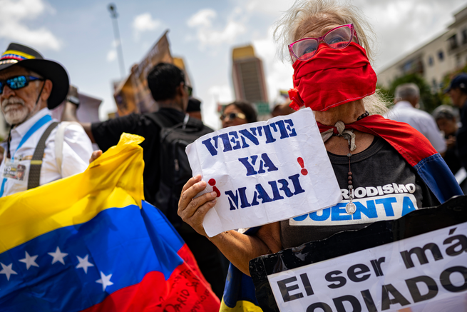 a woman wearing pink-framed glasses and a red face covering holds a sign that says