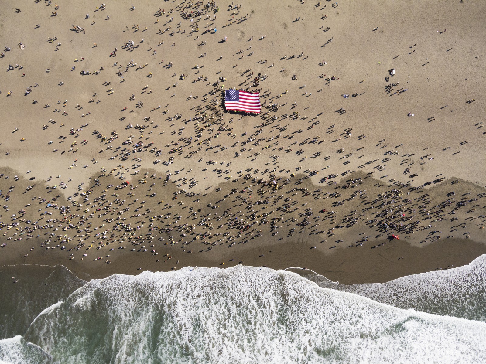 An aerial view of hundreds of people gathered on a beach, many holding up a large American flag.