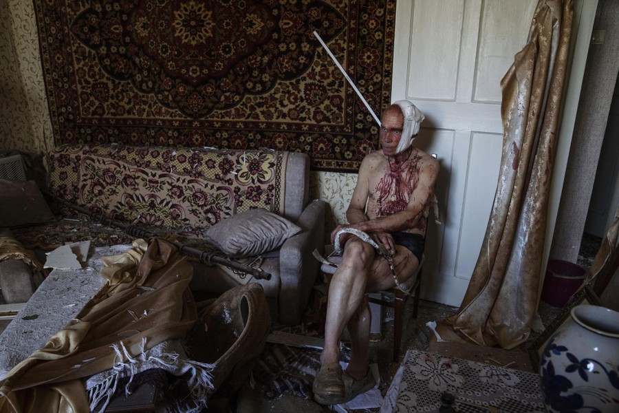 An injured man, his chest covered in blood from an apparent head wound, sits cross-legged in a chair in a damaged apartment.