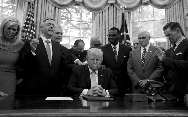 President Trump, Vice President Mike Pence, and faith leaders say a prayer in the Oval Office in September 2017. Several people have their hands on Trump's shoulders. Most of the people have their eyes closed.