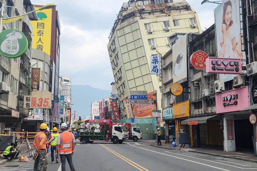 Workers stand in a city street, looking toward a building that is tilting precariously toward the road.