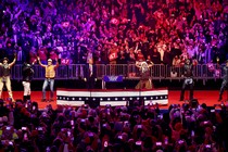 Five men in different costumes dance on a big stage with another man in a suit and red tie.