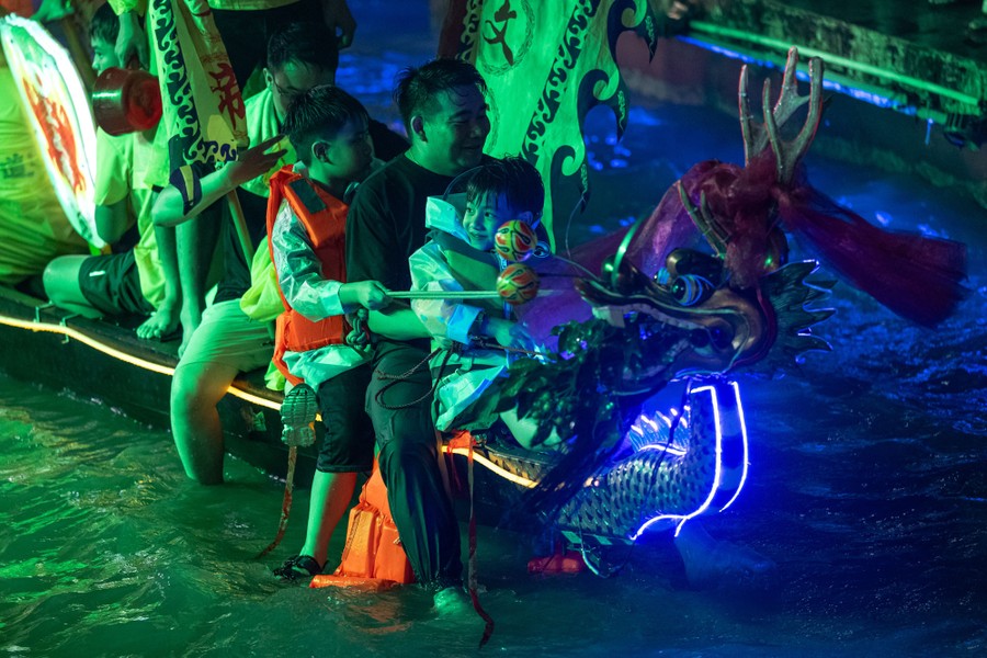 Two adults and two children ride on the front of a neon-lit dragon boat at night.