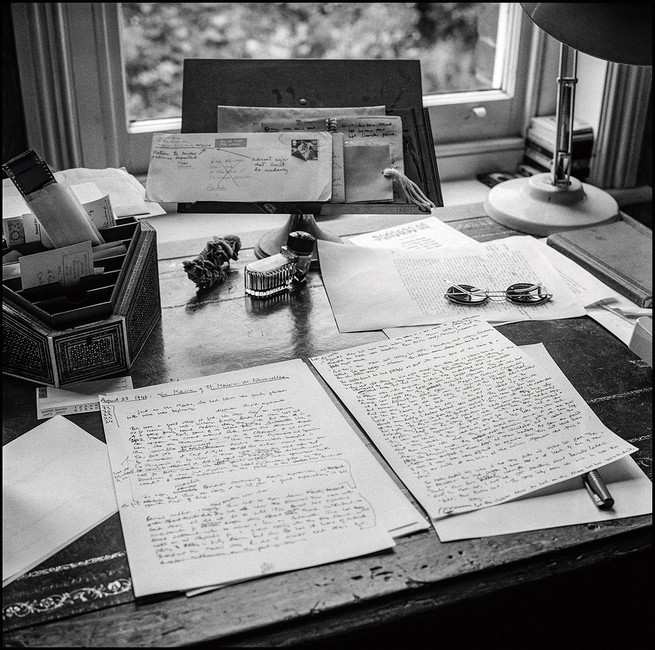 black and white photo of wooden desk by window with handwritten pages, pens, lamp, and correspondence
