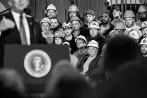Black and white photo of Donald Trump speaking at a podium with workers wearing hard hats seated behind him.