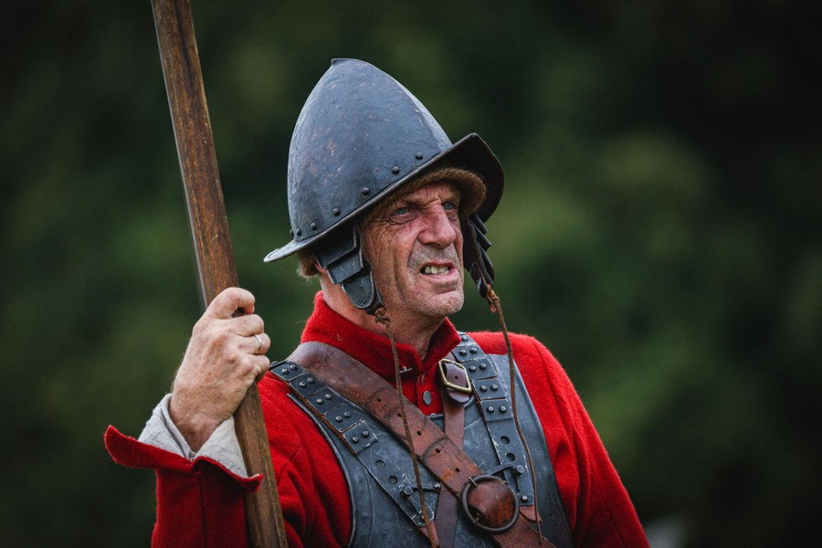 A man in a historical military uniform stands outside, holding a long pike.
