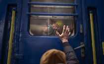 A father stands outside a train, waving goodbye to his wife and young son, who are on the other side of a window, smiling and waving back.