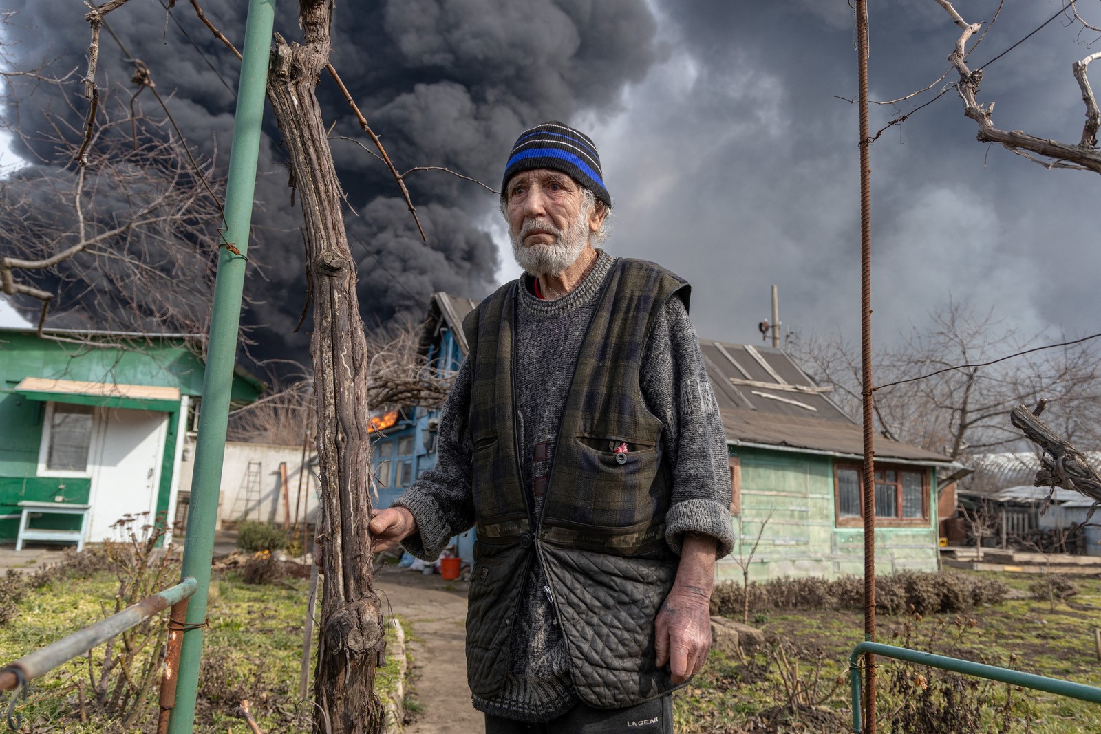 A man stands on a pathway in a courtyard outside a small house, with a large column of black smoke billowing into the air behind him.