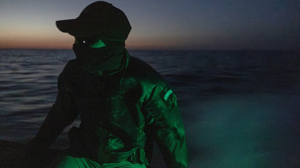 Photograph of a masked soldier on a small boat at dusk, with a green light shining on him