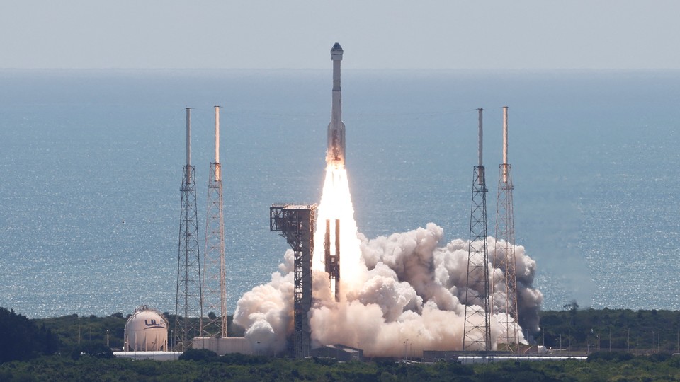 A United Launch Alliance Atlas V rocket carrying two astronauts aboard Boeing’s Starliner-1 Crew Flight Test is launched from the coast of Florida, with blue water creating a backdrop for the flames and the clouds of smoke along the ground.