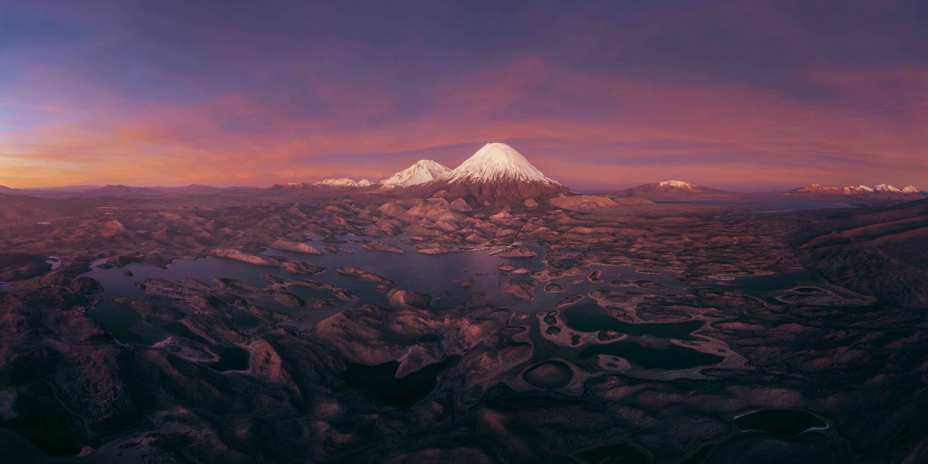 An aerial view of distant snowcapped mountains standing above an uneven field of foothills