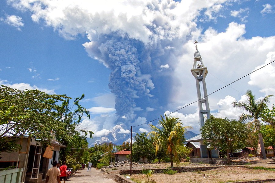 People stand outside a building, looking up toward a distant ash cloud rising from an erupting volcano.