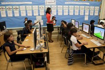 A teacher stands at the back of a computer lab where students are seated at Mac desktop computers.