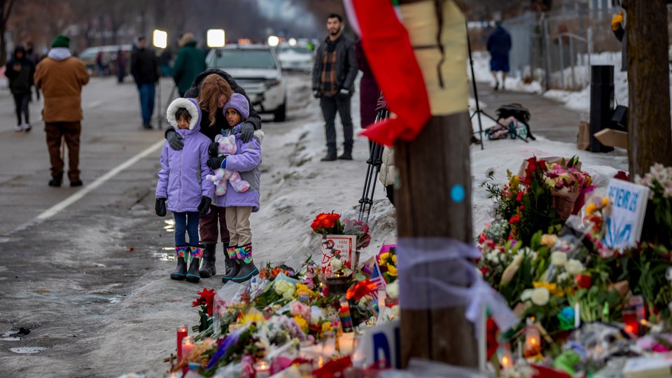 Two little girls in purple coats (one holding a stuffed animal) stand near a flower memorial. Other people are seen in the background.