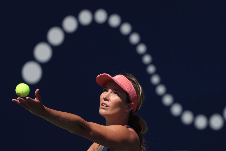 A person serves a tennis ball, tossing it in front of a swooping image of many circles in the background, looking like the ball might be part of the sign.