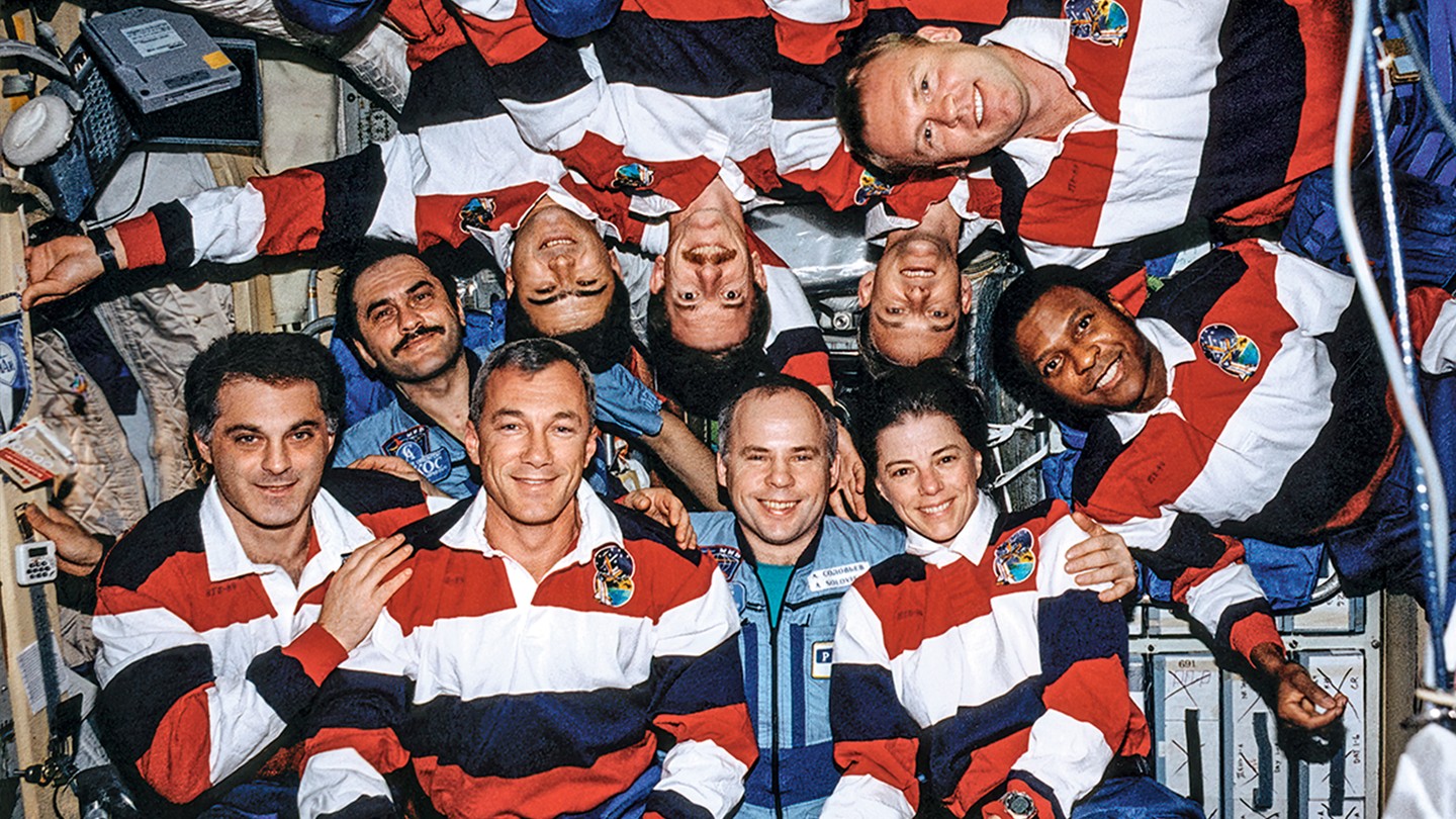 group photo of 10 astronauts in space station, some upside down, most wearing matching red white and blue polo shirts