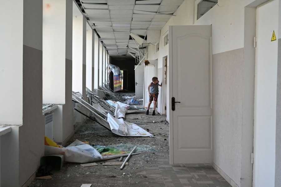 A boy peeks into a classroom in a hallway strewn with broken windows and debris.