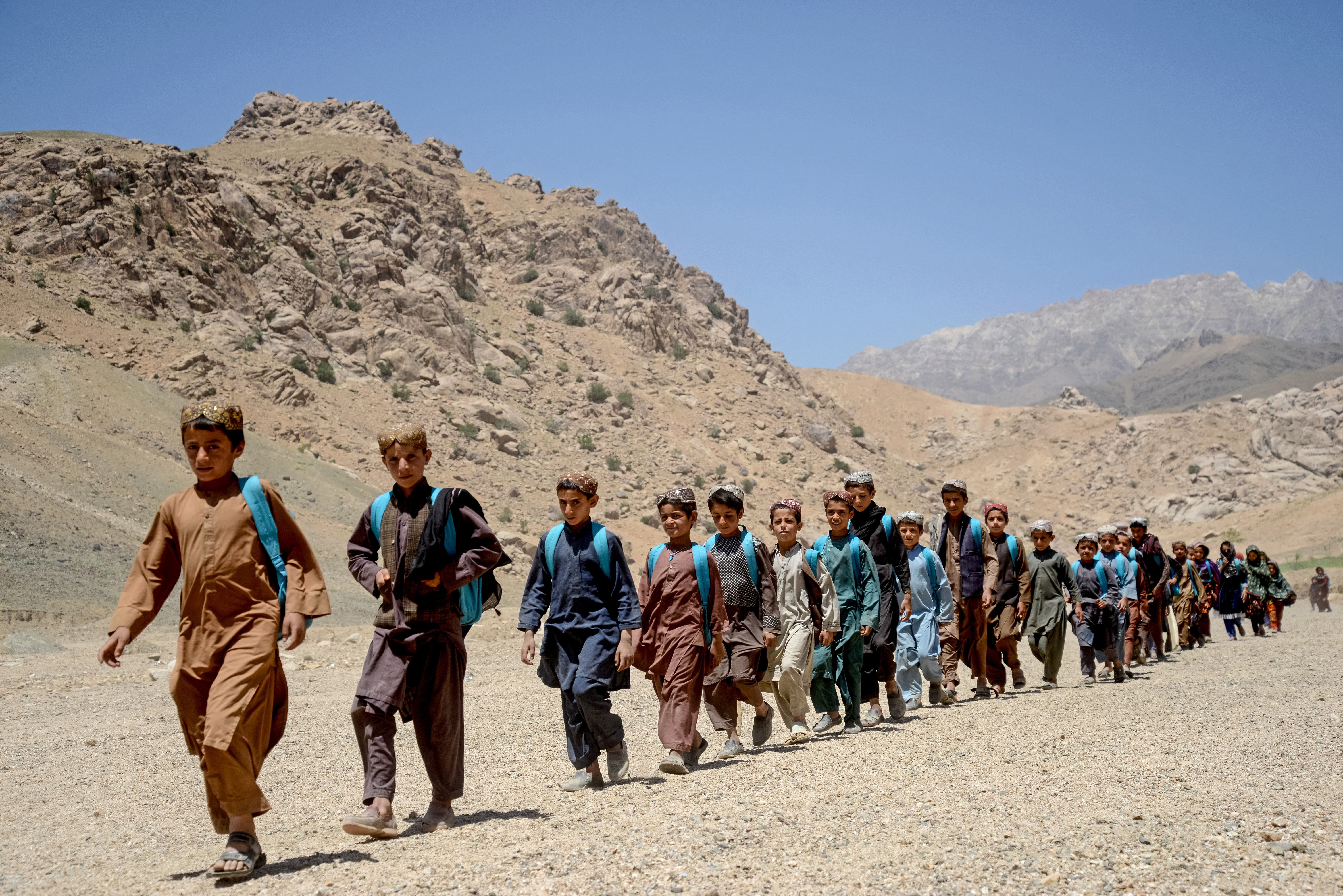 A line of school children with backpacks walk through a dry and mountainous area in Afghanistan.