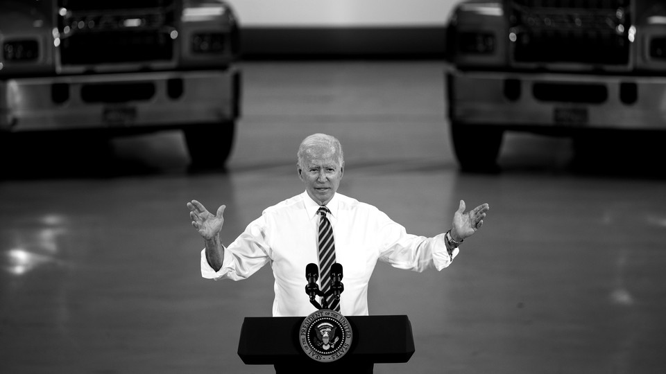 A black-and-white photo of Joe Biden speaking at a podium in front of two trucks.