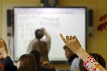 A student raises her hand as her teacher leads a lesson on a SmartBoard. Questions are being raised about whether teachers should be paid for the lessons they create.