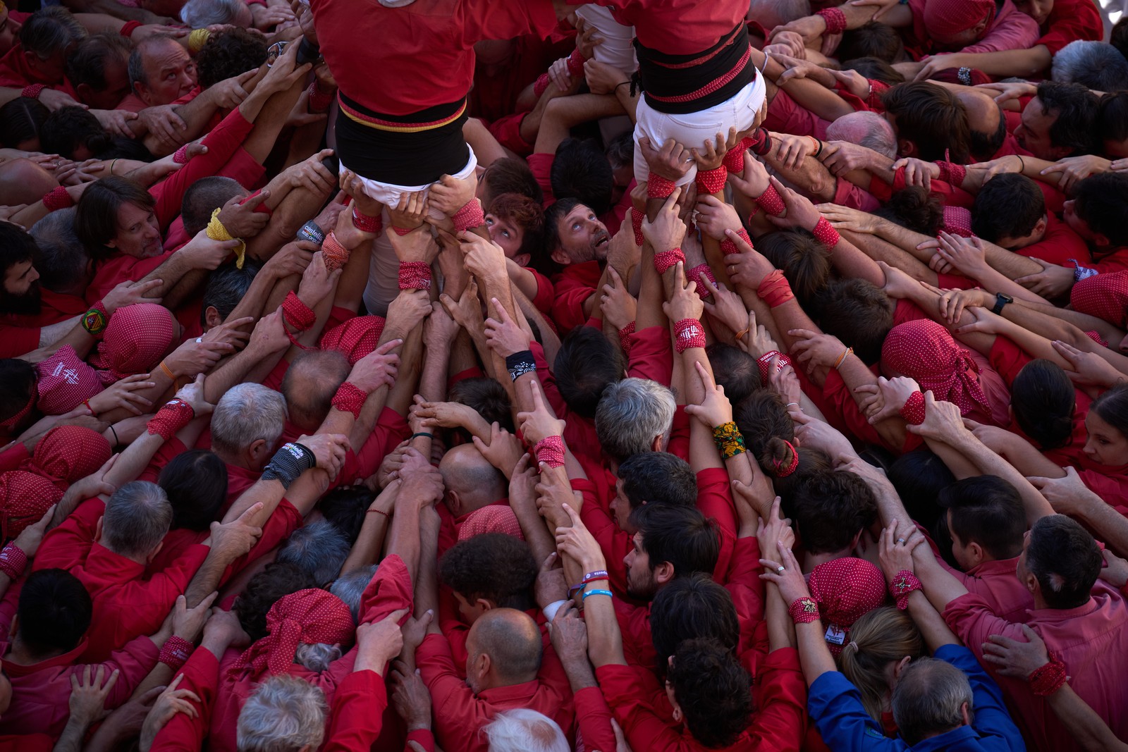 Many people crush together, pushing on each other from behind, working to support several people in the center to form a human tower.