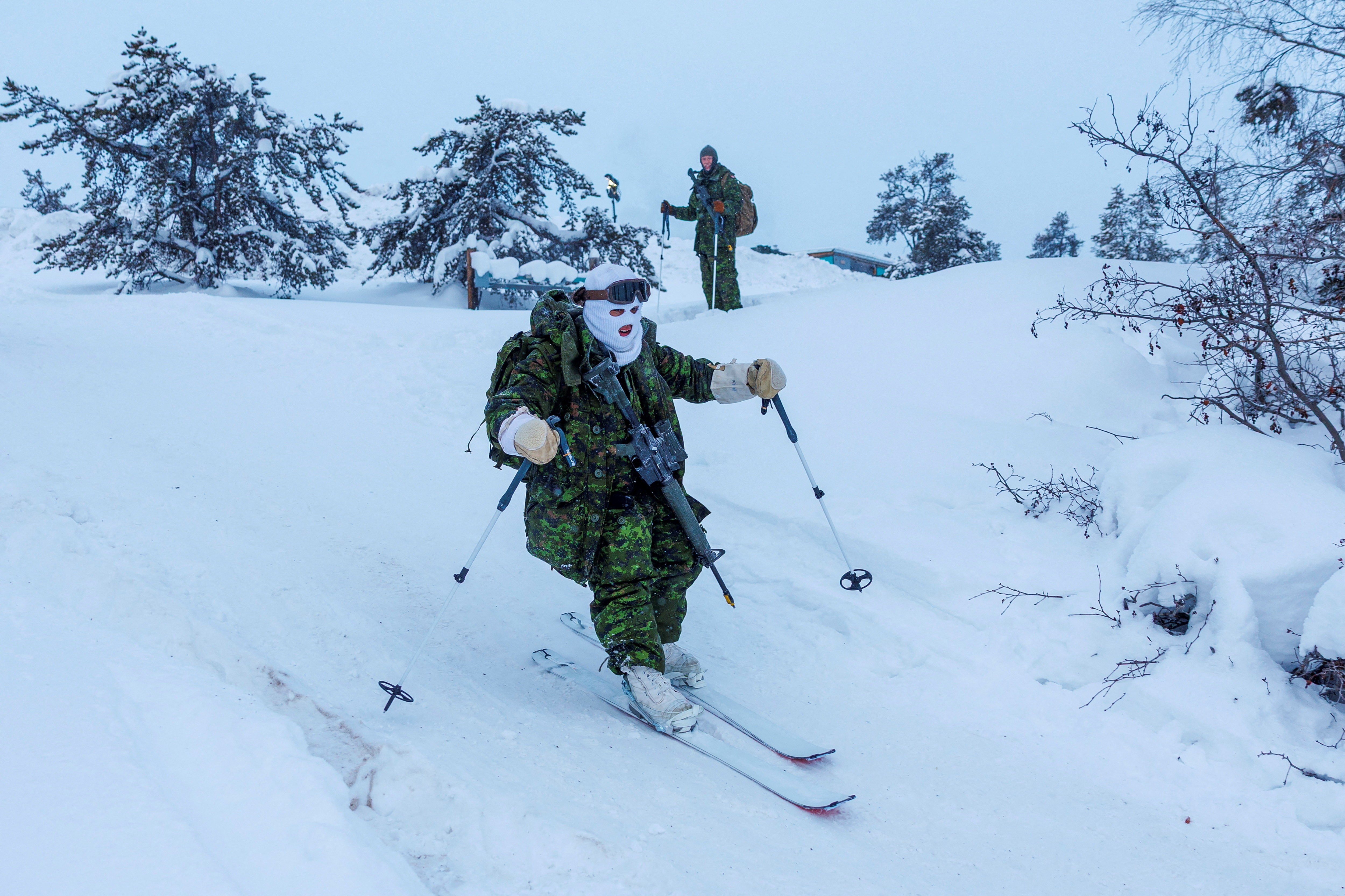 Soldiers wearing cold-weather gear and heavy packs ski across snowy ground.