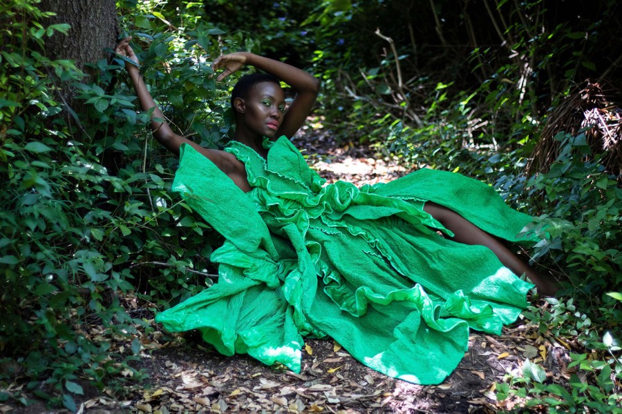 A model poses in a large green gown, among trees and bushes.