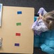 A child and an examiner sit at a table that includes colored blocks, a clipboard, and plastic spoons.