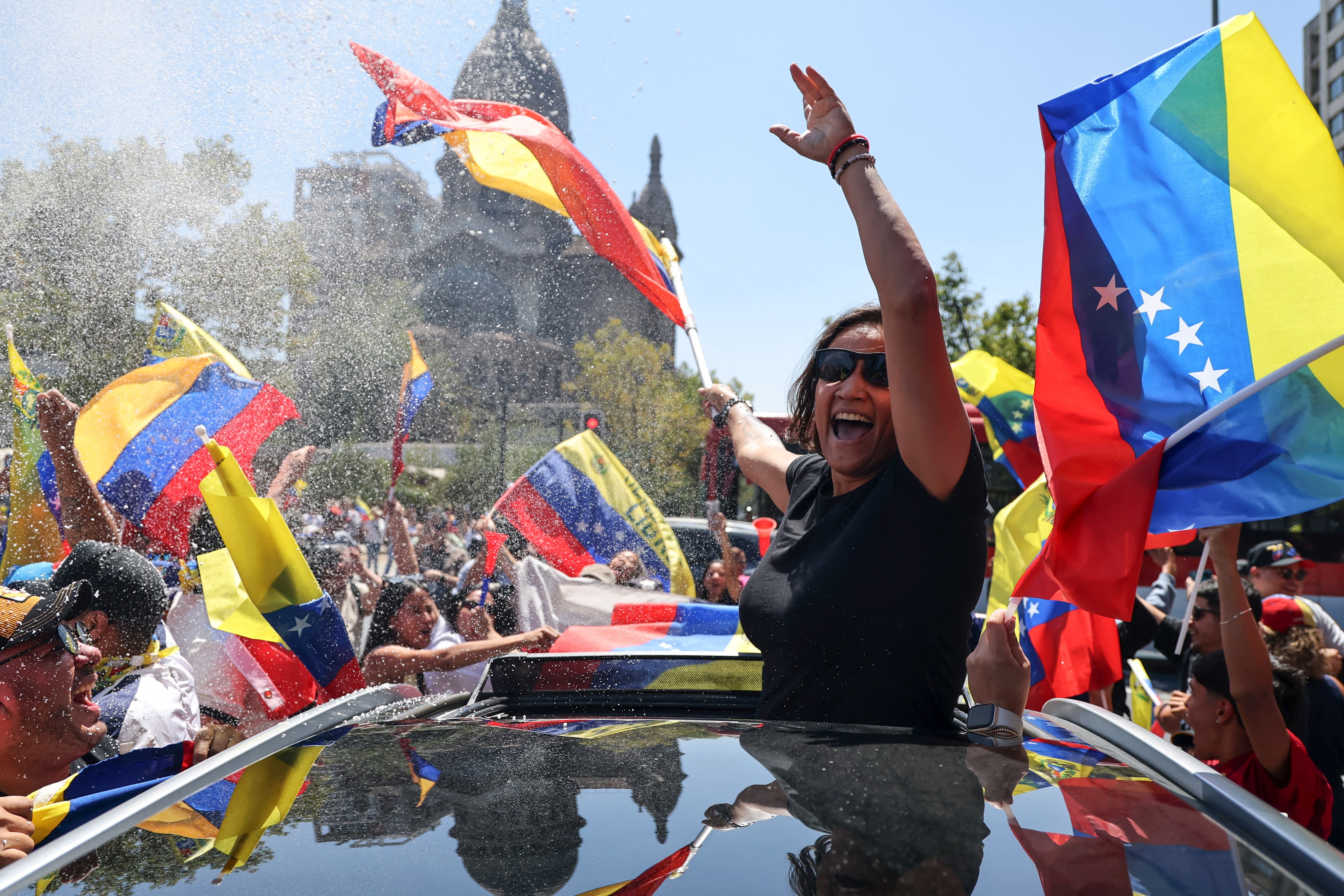A person stands through the open roof of a car, celebrating among a crowd of others, waving Venezuelan flags.