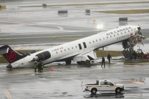 Photograph of the wreckage of Air Canada airplane on the runway of an airport, with the plane’s nose upturned with exposed wiring and torn metal exposing its inside