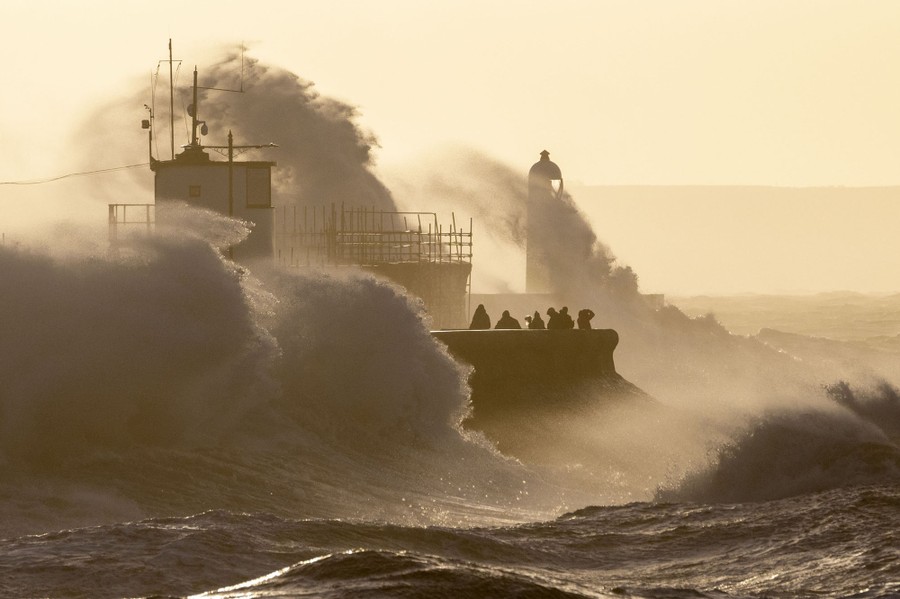 People watch as high waves crash against a harbor wall.