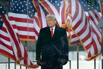 Donald Trump standing outside, behind clear barriers and in front of a row of billowing American flags