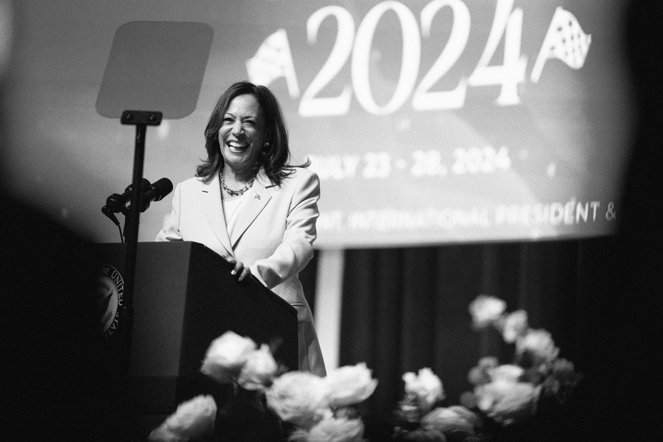 Kamala Harris stands at a podium smiling in front of a large banner that reads 2024