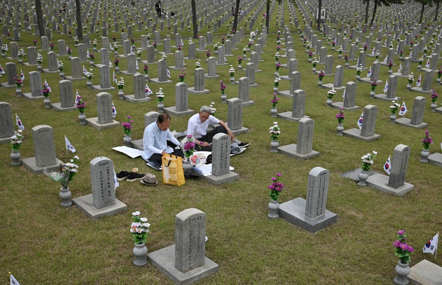 Two people sit before a tombstone in a large cemetery.