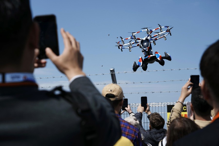 A crowd of people use their phones to take video of a small, one-person helicopter with many small propellers.