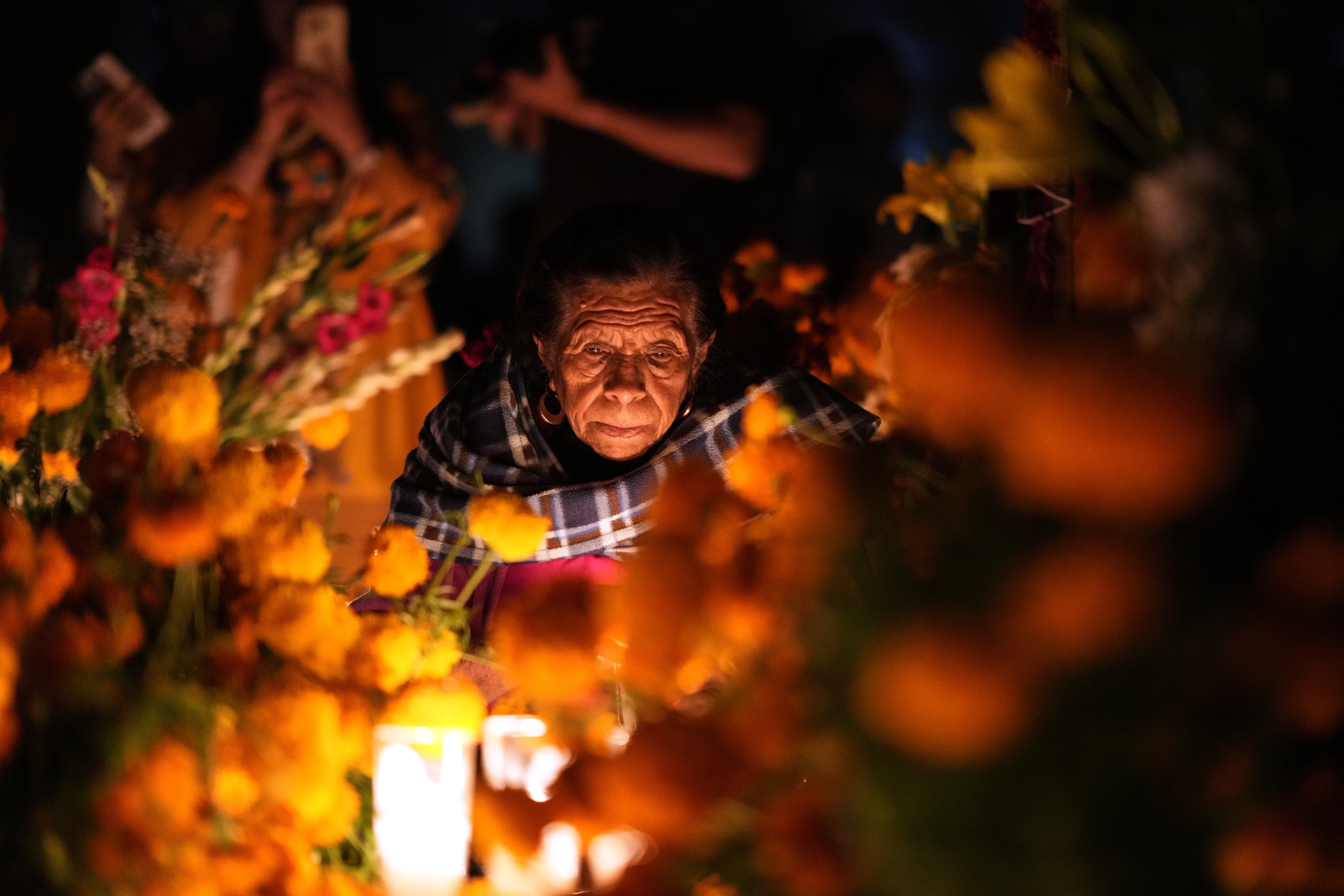A woman leans down next to the grave of a loved one, surrounded by flowers and candles.