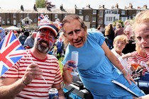 A man wearing Union Jack face paint and a woman wearing a King Charles face mask celebrate the coronation in a field while a child looks on, deeply distraught