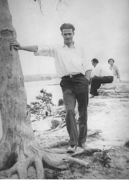 black-and-white photo of standing man, wearing white button-down shirt with sleeves rolled up and trousers, leaning one  hand on a large tree trunk with two figures sitting in distance