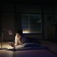 A photograph of a person laying on the floor of a cozy bedroom at night with a lamp illuminating the book the person is reading