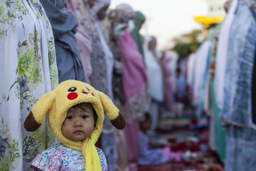 A young child wearing a fuzzy Pikachu hat looks at the camera, as women around her take part in a prayer.