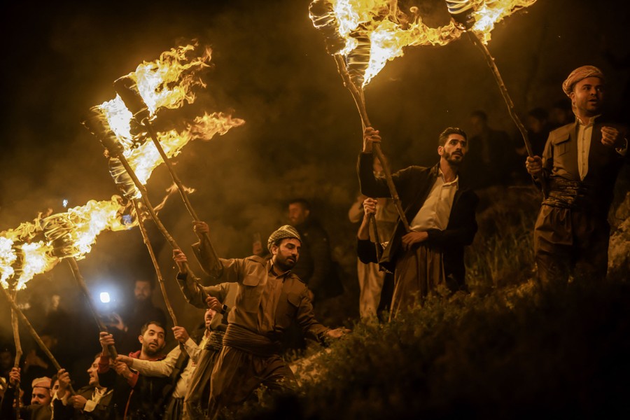 A line of men walk on a mountainside path, carrying large flaming torches.
