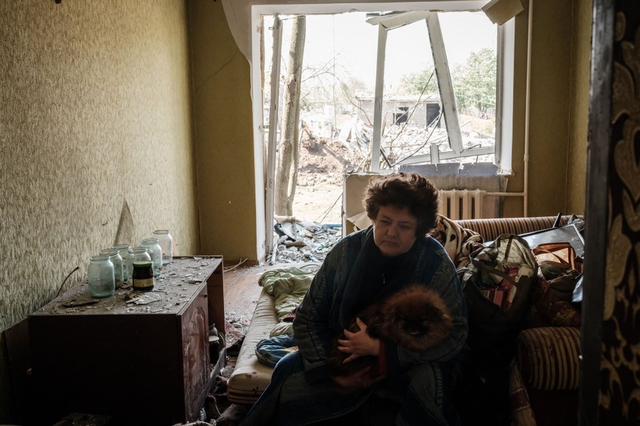 A woman sits on a bed with a small dog in an apartment damaged by a missile explosion.