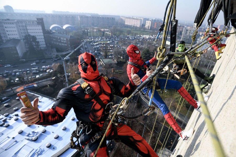 Four people dressed in superhero costumes hang from ropes on the outside of a tall hospital building.