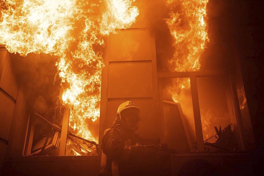 A firefighter holds a hose and stands beside a burning building with huge flames erupting from two windows.