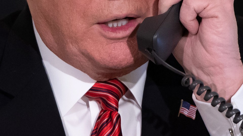 A close-up shot of Donald Trump wearing a black suit jacket, a white shirt, and a red-and-black-striped tie, holding a black phone receiver in front of his mouth