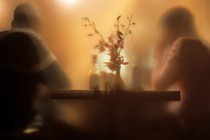 A blurry image of two people seated at a warmly lit table, a vase of flowers between them