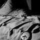 In a black-and-white photo, a watch hangs off an unmade bed cast in shadows.